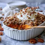 Sweet potato casserole topped with a thick brown sugar pecan crust and a large swirl of whipped cream in a white baking dish.