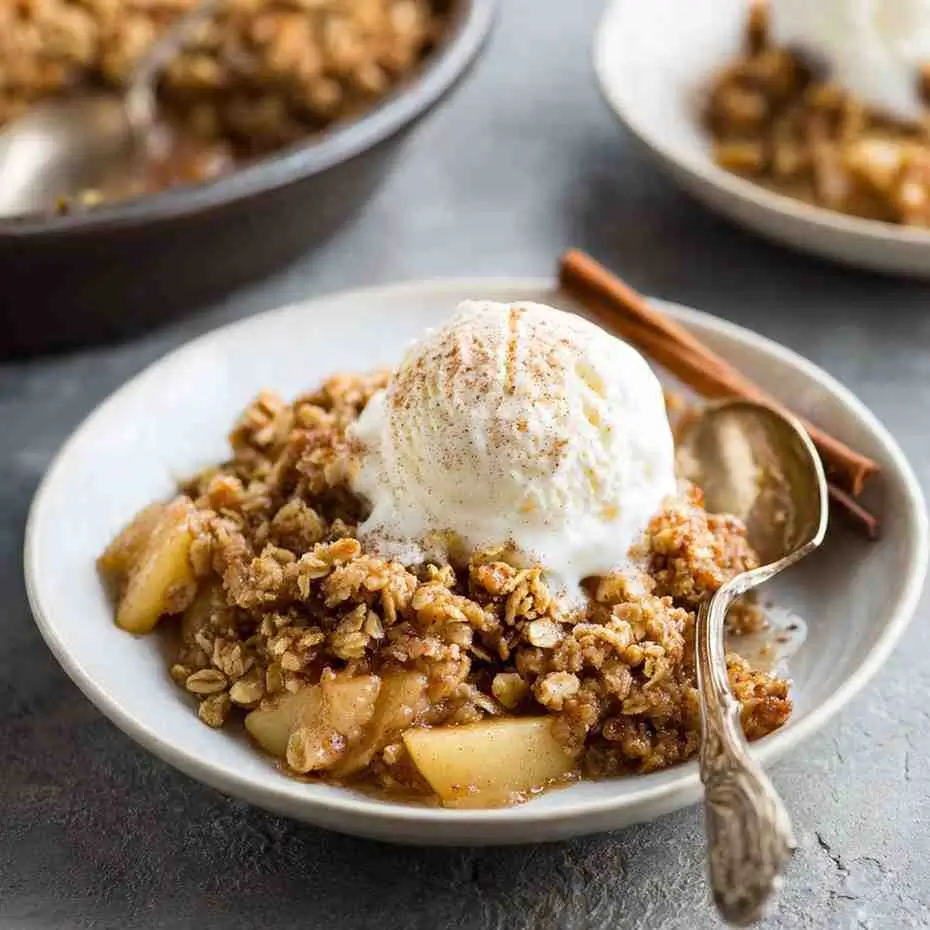 Warm gluten free apple crisp topped with vanilla ice cream and cinnamon, served on a white plate with a spoon.