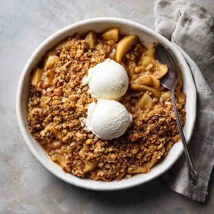 Bowl of maple syrup apple crisp with two scoops of vanilla ice cream on top.