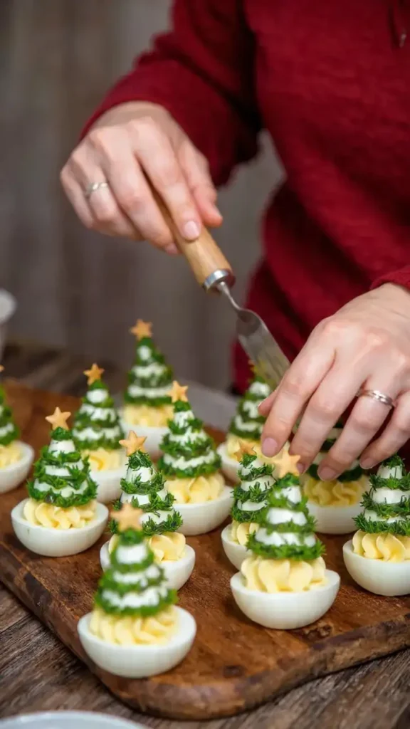 Hands placing herb-covered Christmas tree toppers onto deviled eggs lined on a wooden board.