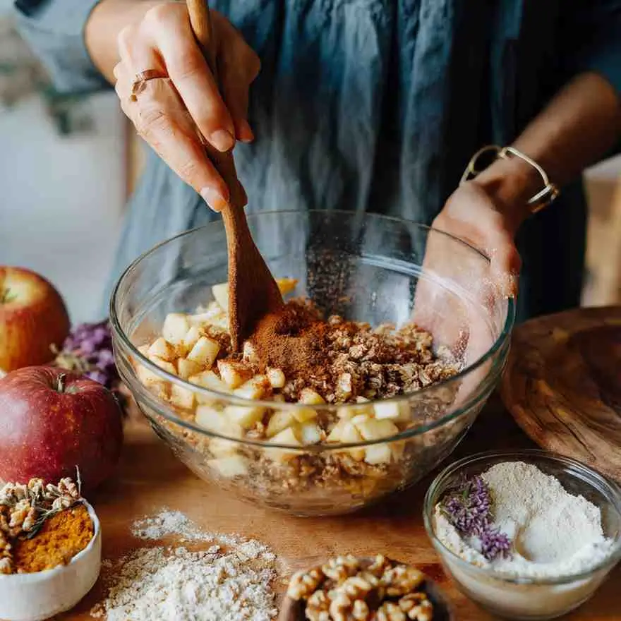 Person stirring chopped apples with cinnamon and oats in a glass bowl for vegan apple crisp.