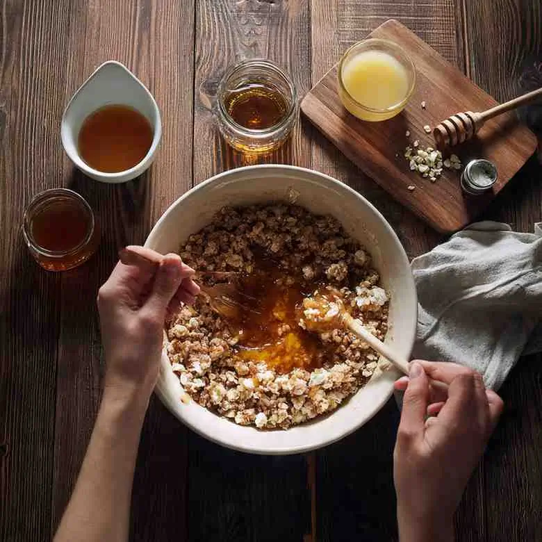 Hands mixing oat crumble with maple syrup in a large bowl on a wooden table.