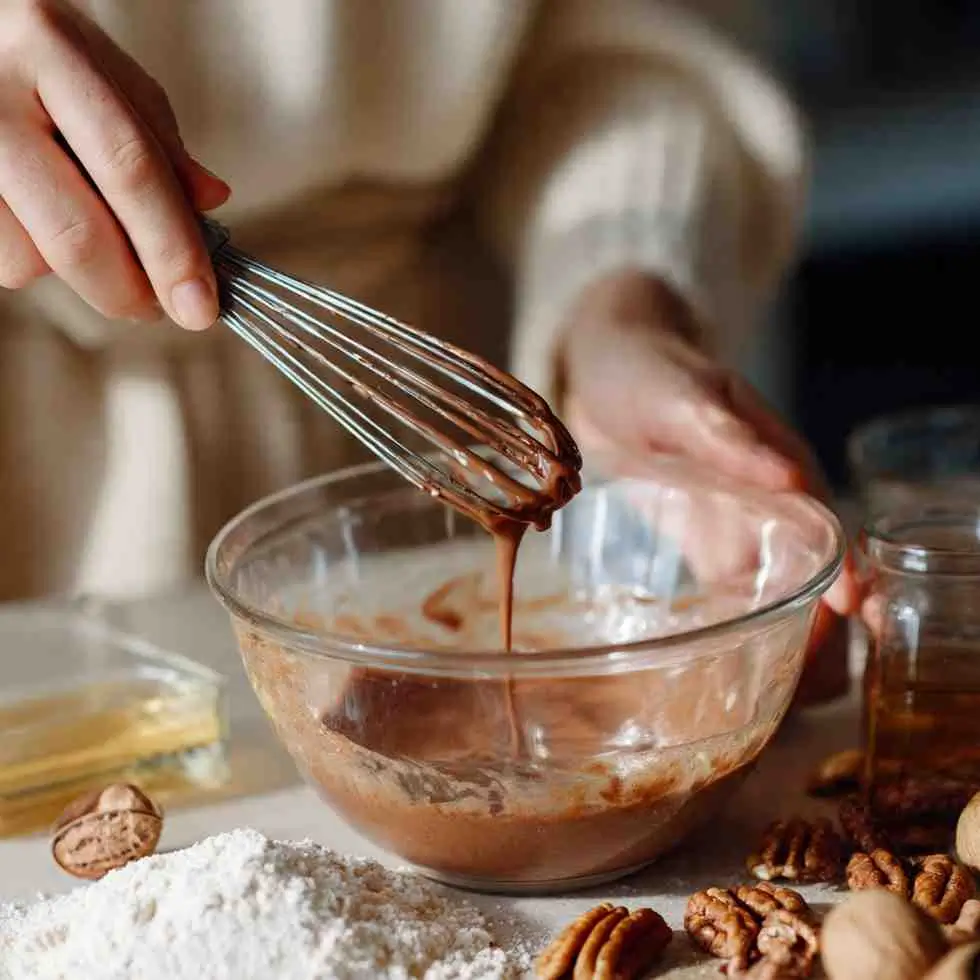 A hand using a wooden spoon to mix flour into a bowl of wet ingredients, surrounded by eggs, nuts, chocolate pieces, and baking supplies.