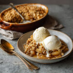 A plate of classic apple crisp topped with two scoops of vanilla ice cream, with a larger baking dish in the background.