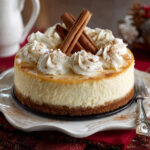 A whole eggnog cheesecake with whipped cream swirls and cinnamon sticks on top, displayed on a white plate.