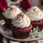 Red velvet cupcakes with tall cream cheese frosting swirls, garnished with red crumbs, with flowers in the background.